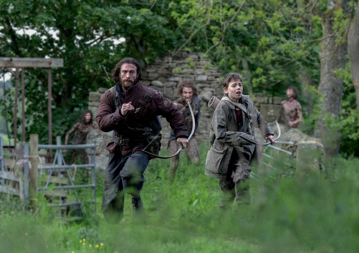 A man in a dark red jacket holding a bow, and a boy in a dark green jacket holding a bow in 28 Years Later. They're running from zombies.