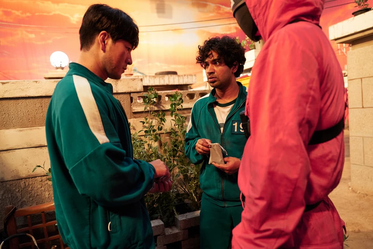 Two men in green jumpsuits standing next to a masked man in a pink suit.