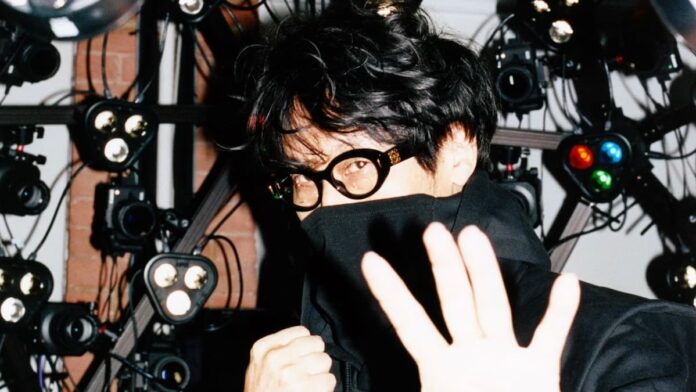 Hideo Kojima for SSENSE Hideo Kojima, a man with dark hair and glasses, dressed in a dark top with his raised towards the camera.