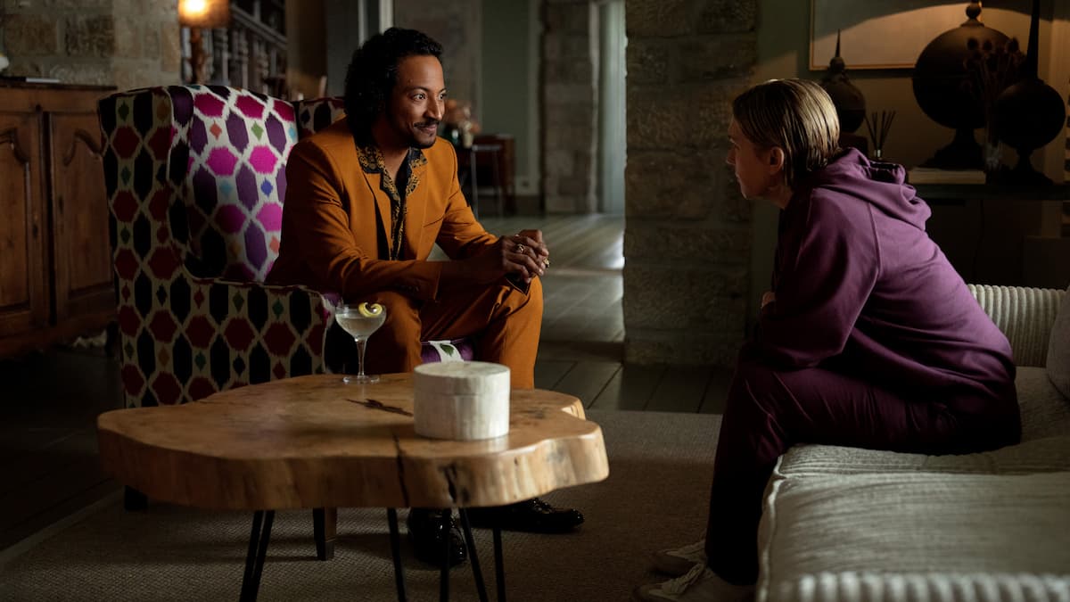 A man in a suit sitting on an armchair talking to a woman in a purple sweatshirt.